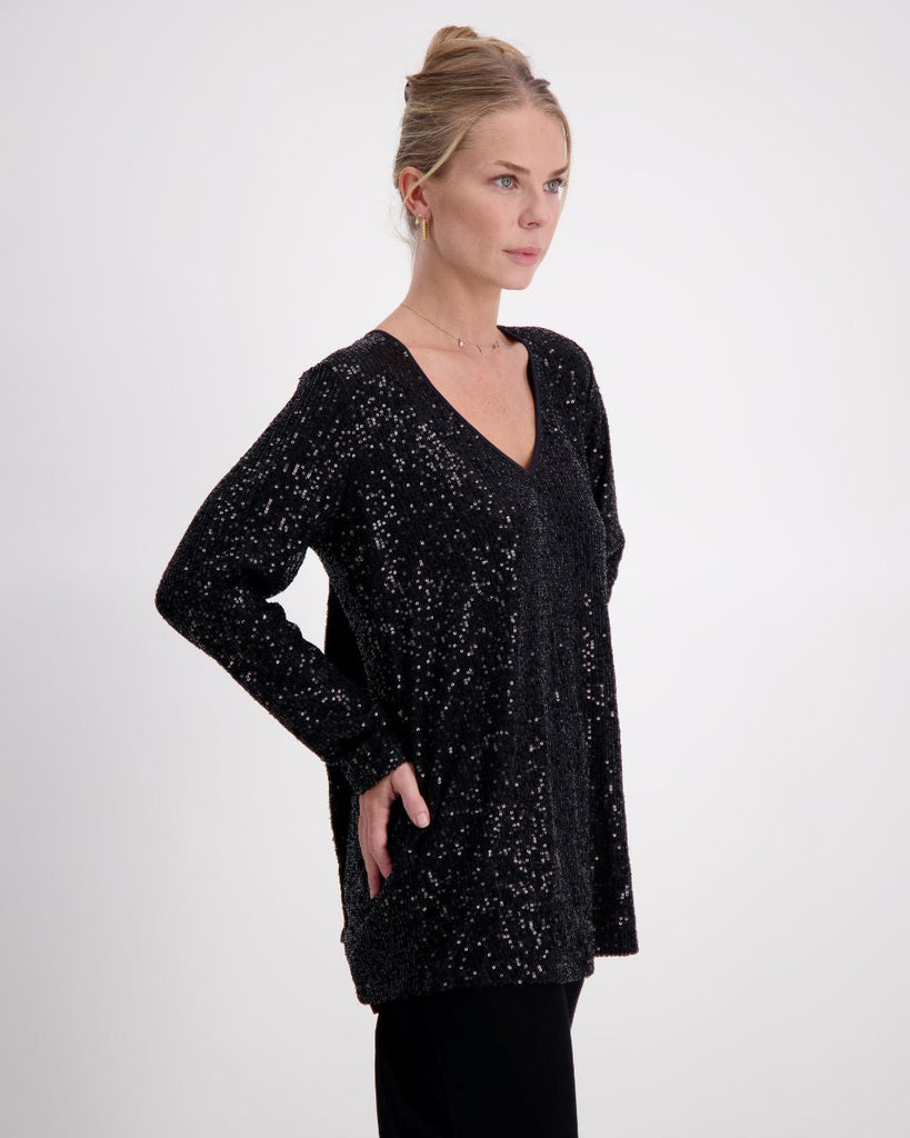 This is a photograph of a woman posing against a white background at a 3/4 angle. She has her hand on her hips. Her blonde hair is styled in an up-do, and she is wearing a black sequinned v-neck top with full length sleeves, paired with black sequin pants. 