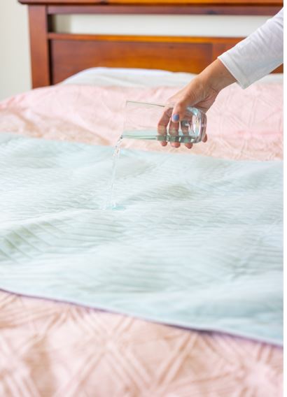 Boss 40 bed pad, on top of a bed with a lady's hand pouring liquid on it to demonstrate its waterproof and absorbent qualities