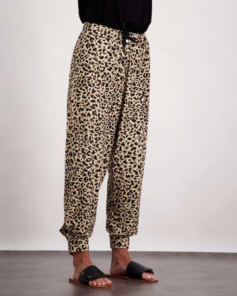 An image of a standing female wearing leopard print slouch pants with a black draw string and black leather slippers. The bottom of her black top is visible. Chistina Stephens Adaptive Clothing Australia. 