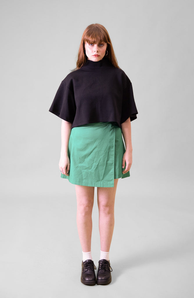 A model standing in front of a grey background wearing a black cropped top and green wrap skirt with black shoes and white socks #Standing